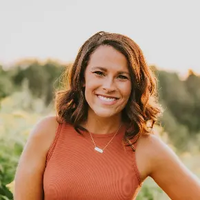 Warm outdoor portrait of a woman, Mrs. Ashley Thomas,  with shoulder-length, wavy brown hair parted to the side, smiling brightly at the camera. She is wearing a rust-orange ribbed tank top and a small silver bar necklace, standing outdoors in front of a softly blurred field with natural sunlight coming from behind.