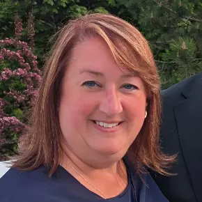 A close-up portrait of Kaye Miziniak, a woman with shoulder-length, strawberry-blonde hair and blue eyes, smiling warmly at the camera. She is wearing a dark navy top, a gold necklace, and small gold earrings. Behind her are lush green trees and vibrant pink flowering bushes outdoors.