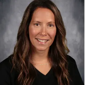 Headshot of a smiling woman, Mrs. Sara Pericolosi, with long brown, wavy hair wearing a black V-neck shirt against a neutral gray studio background.