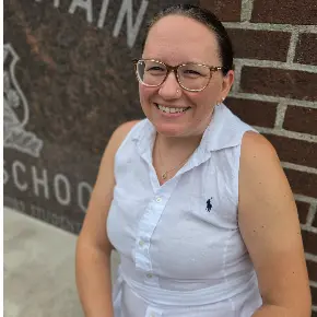 A smiling woman, Mrs. Renee Lindholm,  with her dark hair tied back sits outdoors against a dark brick and granite building wall. She is wearing clear-framed glasses, a gold necklace, and a sleeveless white linen collared shirt featuring a small dark polo player logo. Engraved lettering on the granite section behind her reads "MOUNTAIN" and "SCHOOL".