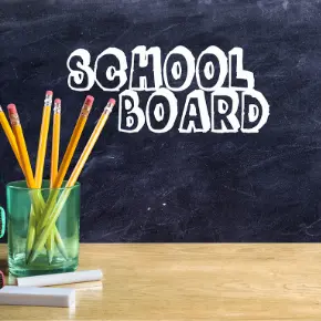"School Board" written on a chalk board with a desk in the foreground featuring pencils in a glass